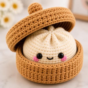 May include: A crocheted cream-colored dumpling with black plastic eyes and pink stitched cheeks, nestled inside a round, tan-colored crocheted steamer basket with a matching lid that is tilted open to reveal the contents inside.