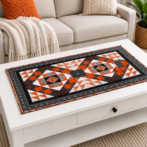 Peut inclure: Un chemin de table matelassé rectangulaire présentant un motif patchwork géométrique en noir, blanc et orange. Le design se compose de motifs en forme d'étoiles et de triangles, entouré d'une bordure en tissu noir avec un imprimé blanc discret.