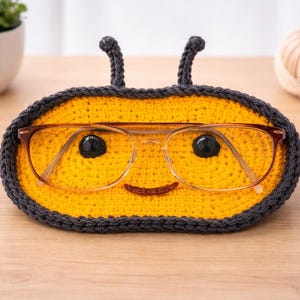 May include: A handmade, yellow and gray crochet glasses holder shaped like a bee. The holder features a smiling face and antennae, with a pair of eyeglasses resting inside. The holder is designed to keep glasses safe and accessible.