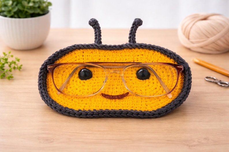 May include: A handmade, yellow and gray crochet glasses holder shaped like a bee. The holder features a smiling face and antennae, with a pair of eyeglasses resting inside. The holder is designed to keep glasses safe and accessible.