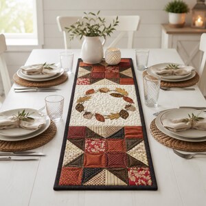 May include: A rectangular quilted table runner featuring a central wreath of autumn leaves on a cream background, flanked by geometric star patterns in shades of red, brown, and tan. The runner is centered on a white dining table with place settings.