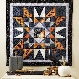 May include: A square wall quilt featuring a central star pattern in black, orange, and white with crow and pumpkin motifs. The quilt has a black border with small yellow dots and intricate stitching. It is displayed with a black bird figurine.