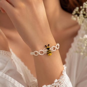 May include: A white crocheted bracelet featuring a series of interconnected circular loops and a central bee motif. The bee is crafted with yellow and black thread, showcasing detailed wings and a striped body, integrated into the delicate lace-like band.