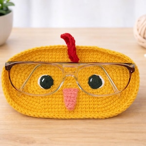 May include: A yellow crocheted eyeglass holder shaped like a chick, with black eyes, a pink beak, and a red comb. The holder contains a pair of eyeglasses. The holder is on a wooden surface.