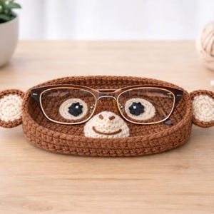 May include: A brown crochet eyeglass holder shaped like a monkey face, with round ears and a smiling mouth. The holder contains a pair of eyeglasses. The holder is on a wooden surface.