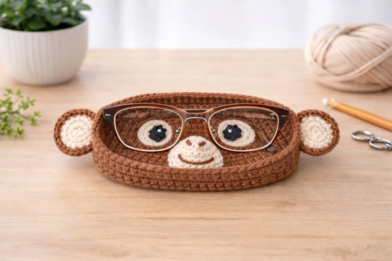 May include: A brown crochet eyeglass holder shaped like a monkey face, with round ears and a smiling mouth. The holder contains a pair of eyeglasses. The holder is on a wooden surface.
