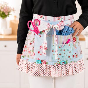 Op de afbeelding: Een schort met bloemenprint met meerdere zakken, een geplooid zoom en een strik. De schort heeft een roze schaar, een blauwe meetlint en andere naai-instrumenten in de zakken. Het ontwerp van de schort bevat rode rozen en stippen.