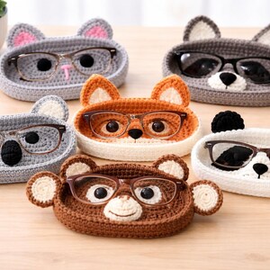 May include: A collection of handmade crochet eyeglass holders in the shape of various animals, including a cat, fox, koala, bear, and panda. Each holder is a different color and holds a pair of eyeglasses. The holders are displayed on a wooden surface.