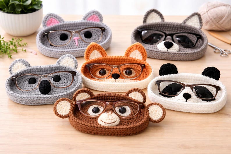 May include: A collection of handmade crochet eyeglass holders in the shape of various animals, including a cat, fox, koala, bear, and panda. Each holder is a different color and holds a pair of eyeglasses. The holders are displayed on a wooden surface.