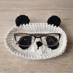 May include: A white and black crochet panda-shaped eyeglass holder with black ears and facial features. The holder contains a pair of black-framed eyeglasses with clear lenses. The holder is on a light brown wooden surface.