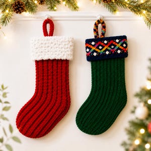 Op de afbeelding: Twee gehaakte kerstsokken hangen naast elkaar. De linker sok is effen rood met een witte boord met textuur en een rode lus. De rechter sok is donkergroen met een marineblauwe boord voorzien van een kleurrijk regenboogpatroon met ruiten.