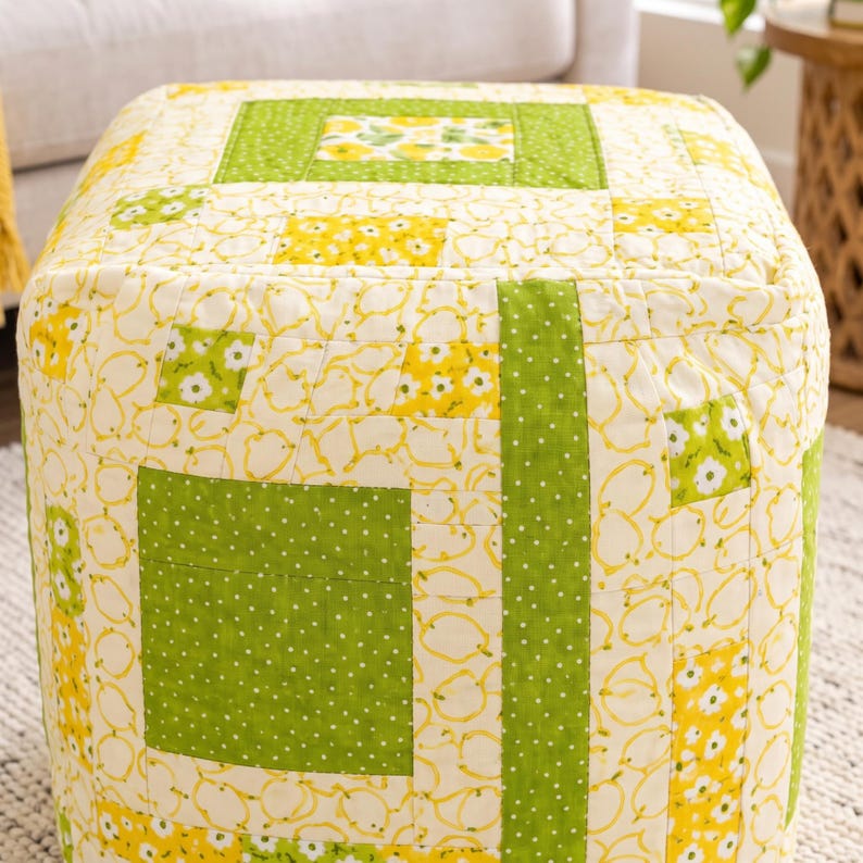 May include: A square ottoman upholstered in a quilted fabric featuring a patchwork design. The pattern consists of alternating blocks of lime green fabric with white polka dots and cream fabric printed with yellow lemons and white flowers.