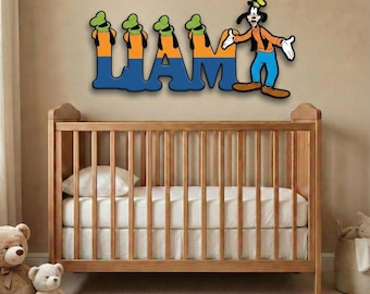 May include: A wooden crib with a white mattress and bedding. Above the crib, the name "LIAM" is displayed in blue letters with cartoon character accents. The cartoon character Goofy is also featured in the design.