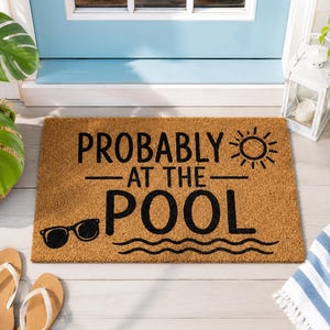 Könnte beinhalten: Eine rechteckige, hellbraune Fußmatte aus Kokosfaser mit dem schwarzen Schriftzug PROBABLY AT THE POOL. Das Design enthält eine Grafik einer Sonne mit Strahlen, eine schwarze Sonnenbrille und drei Wellenlinien, die Wasser darstellen.