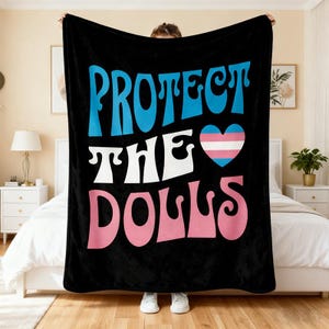May include: Black blanket with the words "PROTECT THE DOLLS" in blue, white, and pink. A heart with the transgender flag colors is in the center. The blanket is held up by a person.