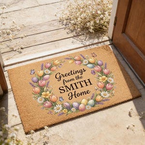 May include: A rectangular doormat with a floral wreath design. The wreath features tulips, daisies, and lavender in shades of pink, yellow, and purple, surrounding the text "Greetings from the SMITH Home". Butterflies and Easter eggs are also included.