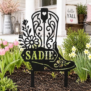 May include: A black metal garden stake featuring a cutout design of a cowboy boot adorned with floral patterns and a heart shape at the top. The name SADIE is cut out in bold capital letters across the center of the boot, supported by two prongs.