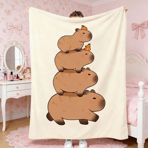 May include: A beige blanket featuring a stack of four cartoon capybaras, each with a small pumpkin on its head. The capybaras are brown with light brown accents. The blanket is held up by a person.