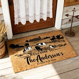 Könnte beinhalten: Eine rechteckige, beigefarbene Fußmatte aus Kokosfaser mit einem schwarzen Aufdruck, der eine Farm mit Kühen auf einer Weide und eine Scheune zeigt. Der Text lautet: WELCOME TO The Anderson's It's Good To Be Home. Die Matte hat eine grobe Struktur.