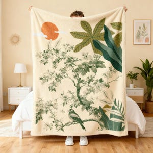 May include: Cream-colored blanket with a botanical design featuring a tree, bird, and foliage in shades of green and white. An orange sun and clouds are also depicted. The blanket is held up by a person.
