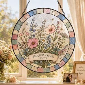 May include: Round stained glass window art featuring a floral design with pink, blue, and white flowers. The design includes a banner with the text "[COUPLE NAMES]" and "[DATE]", and is framed with a colorful border. It hangs from a black chain.