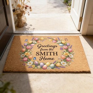 May include: A rectangular coir doormat with a floral wreath design. The wreath features colourful tulips, daisies, and Easter eggs, surrounding the text "Greetings from the SMITH Home". The mat is placed in a doorway.