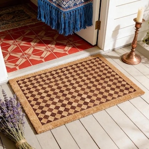 May include: A rectangular doormat with a brown and tan checkered pattern. The mat has a natural fiber appearance and a border. A bundle of lavender sits to the left of the mat. The mat is on a wooden floor.