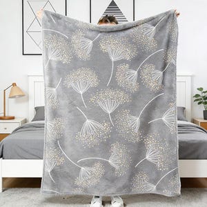 May include: A gray plush blanket with a white floral pattern. The blanket is rectangular and features a repeating design of stylized flowers with dotted centers and thin stems. The blanket is held up in front of a bed.