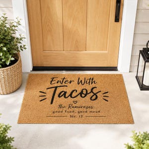 Könnte beinhalten: Eine rechteckige Fußmatte aus natürlicher Kokosfaser. Die Matte trägt den Schriftzug "Enter With Tacos" in schwarzer Schrift sowie zusätzlichen Text. Die Matte liegt vor einer Holztür.