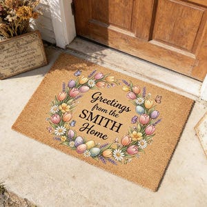May include: A rectangular coir doormat with a floral wreath design. The wreath features tulips, daisies, and Easter eggs, surrounding the text "Greetings from the SMITH Home". The mat is placed in front of a wooden door.