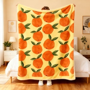 May include: A yellow blanket with a repeating pattern of orange fruit with green leaves. The blanket is held up, showcasing the design. The blanket is a cozy home decor item.