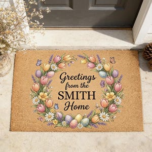 May include: A rectangular coir doormat with a floral wreath design. The wreath features tulips, daisies, and lavender, surrounding the text "Greetings from the SMITH Home." Easter eggs and butterflies are also included in the design.