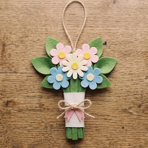 May include: A felt flower bouquet ornament featuring pink, white, and blue flowers with yellow centers and green leaves. The stems are wrapped in a white band with a small pink bow and a jute twine loop at the top for hanging the decoration.