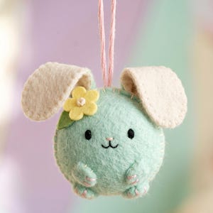 May include: A round, mint green felt ornament shaped like a rabbit with long cream-colored ears, a small yellow felt flower on its head, and embroidered black eyes and mouth. The plush figure hangs from a thin pink string attached to the top.