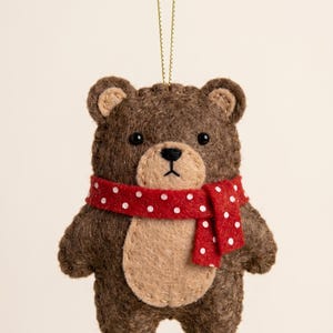 May include: A brown felt bear ornament featuring a tan belly patch, black bead eyes, and a stitched nose. The bear wears a red scarf with white polka dots and is suspended by a thin gold string for hanging. The edges are finished with visible stitching.
