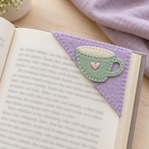 May include: A triangular felt corner bookmark in light purple featuring a stitched green tea cup applique with a cream-colored top and a small pink embroidered heart in the center, placed on the corner of an open book page.