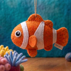 May include: A felt clownfish ornament in vibrant orange with white vertical stripes and a black-rimmed eye. The fish hangs from a thin twine string and features a small white tag with the text TINIMALS attached to its side fin area.
