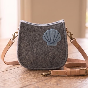 May include: A grey felt crossbody bag with a light blue seashell applique on the front. The bag features a tan leather adjustable shoulder strap attached with brass-toned metal swivel clips and rivets, creating a structured, compact accessory.