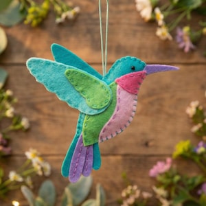 May include: A vibrant felt hummingbird decoration with turquoise wings, a green back, and a pink throat. The bird features a purple beak and tail feathers. It hangs by a string against a wooden backdrop with foliage and fairy lights.