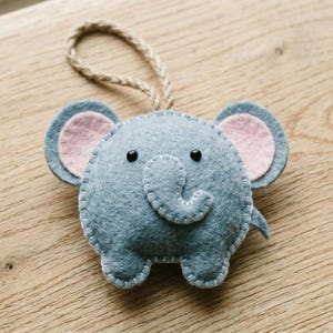 Puede incluir: Un adorno de fieltro gris con forma de elefante, orejas rosadas, dos pequeños ojos de cuentas negras y una trompa curvada. La figura está rematada con puntadas de festón blancas e incluye un lazo de cuerda de yute para colgar el objeto.