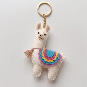 Puede incluir: Un llavero de fieltro color crema con forma de llama y una anilla con cadena de metal dorado. La llama tiene ojos y boca bordados en negro, con una manta de silla de montar colorida en capas de fieltro azul, amarillo, rosa y morado y una borla.