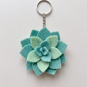May include: A felt succulent keychain featuring layered petals in alternating shades of teal and light mint green. The plant-shaped charm is attached to a silver-toned metal split ring by a short chain, creating a floral design with a soft texture.