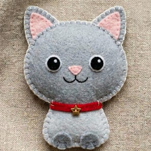 Peut inclure: Un ornement en feutrine grise représentant un chat avec des oreilles roses à l'intérieur, de grands yeux noirs, un nez rose et une bouche brodée. Le chat porte un collier rouge avec une petite étoile dorée au centre et des bords cousus.