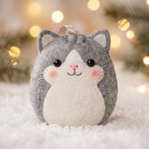 Pode incluir: Enfeite de gato de feltro cinza e branco com um rosto sorridente, bochechas rosadas e nariz rosa. O gato tem uma barriga branca e uma alça para pendurar. O fundo inclui uma superfície nevada e luzes de Natal desfocadas.