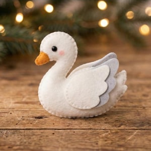 May include: A handmade white felt swan with a curved neck, orange beak, and grey wing details. The swan has a sweet expression with pink cheeks and black eyes. It is placed on a wooden surface with blurred lights in the background.
