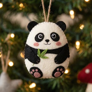 May include: A felt panda bear Christmas decoration. The ornament is white with black detailing, including the ears, arms, and legs. It has a smiling face with pink cheeks and holds a green leaf. Other festive decorations are in the background.