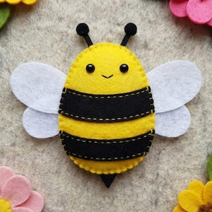 May include: A handmade felt bee with yellow and black stripes, white wings, and black antennae. The bee is surrounded by felt flowers and green leaves in shades of pink, yellow, and green. The background is a light gray.