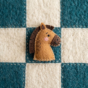 May include: A tan felt horse head ornament with a dark brown mane and muzzle, featuring embroidered black eyes, a stitched nose, and pink circular cheeks. The horse is positioned on a teal and cream checkered felt background with white stitching.