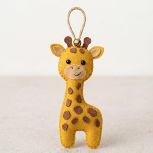 May include: A yellow felt giraffe ornament featuring brown spots, black bead eyes, and a stitched smile. The giraffe has two small brown horns and a beige muzzle. A gold-coloured metal bell is attached to the top, connected to a beige rope loop.