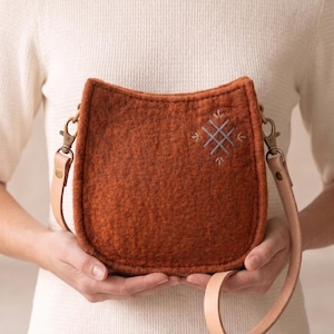 May include: A rust-colored felted wool crossbody bag with a curved top edge and a light tan leather strap attached by brass-toned metal swivel clips. The front of the bag features a small, embroidered geometric cross pattern in light gray thread.